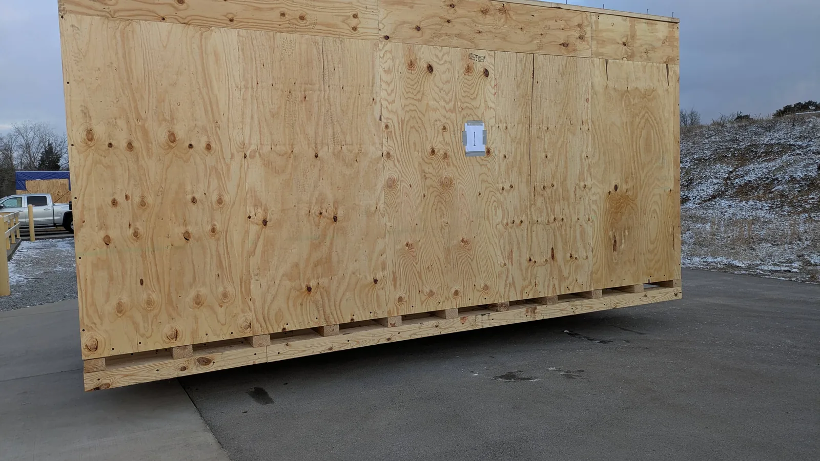 A large wooden export crate on a forklift-accessible base, outdoors beside a pickup truck.