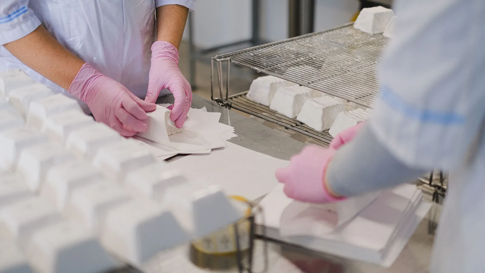 Workers in gloves wrapping product by hand at a stainless-steel bench.